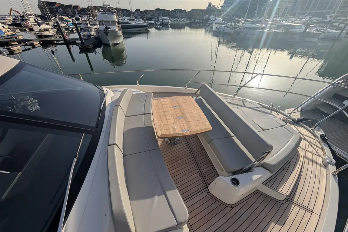 An elegant yacht's foredeck with grey seating, a wooden table, and a serene marina view reflecting the sunlight.